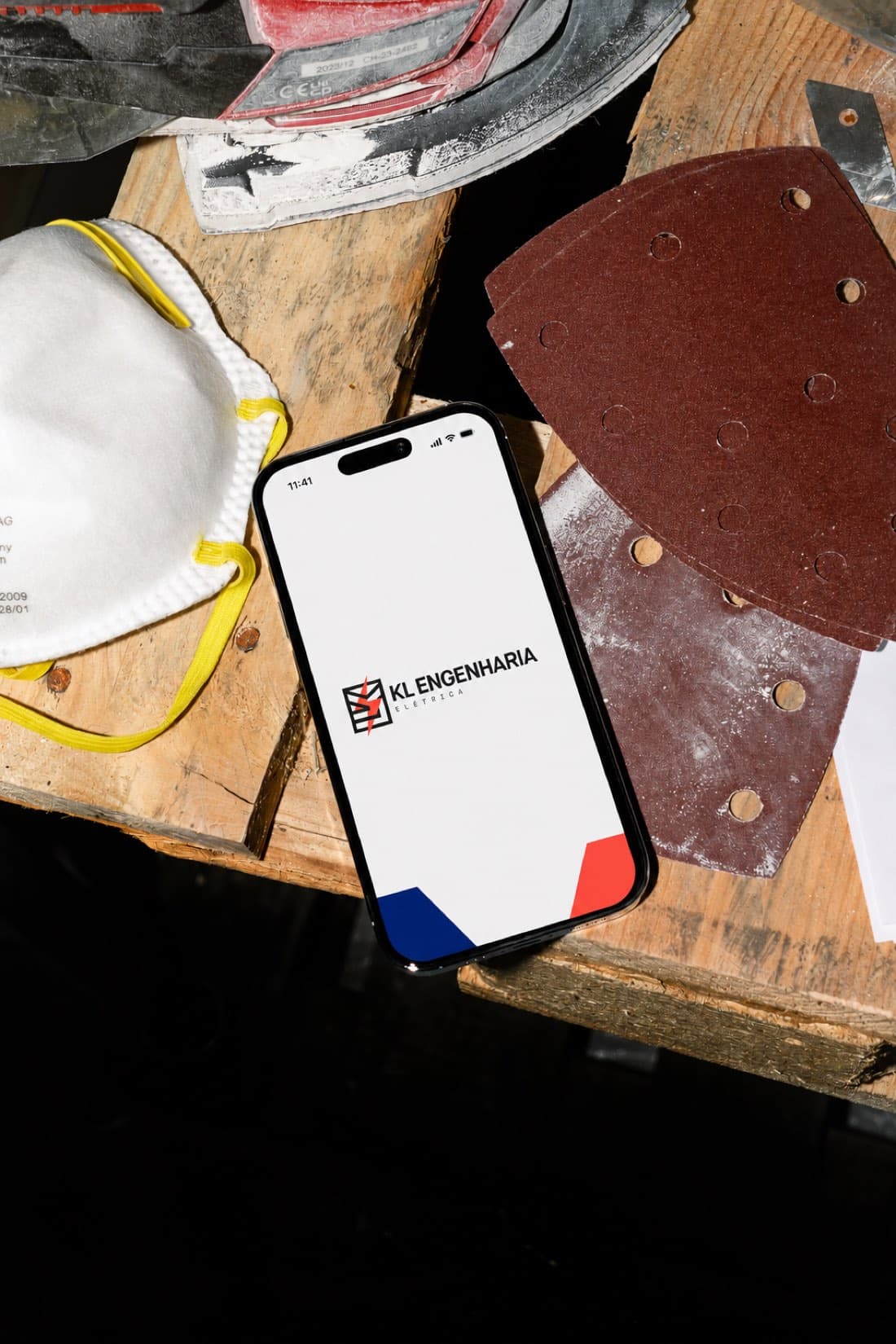 Novo site da KL Engenharia Elétrica aberto em um celular apoiado na bancada de obra, entre máscara e discos de lixa.